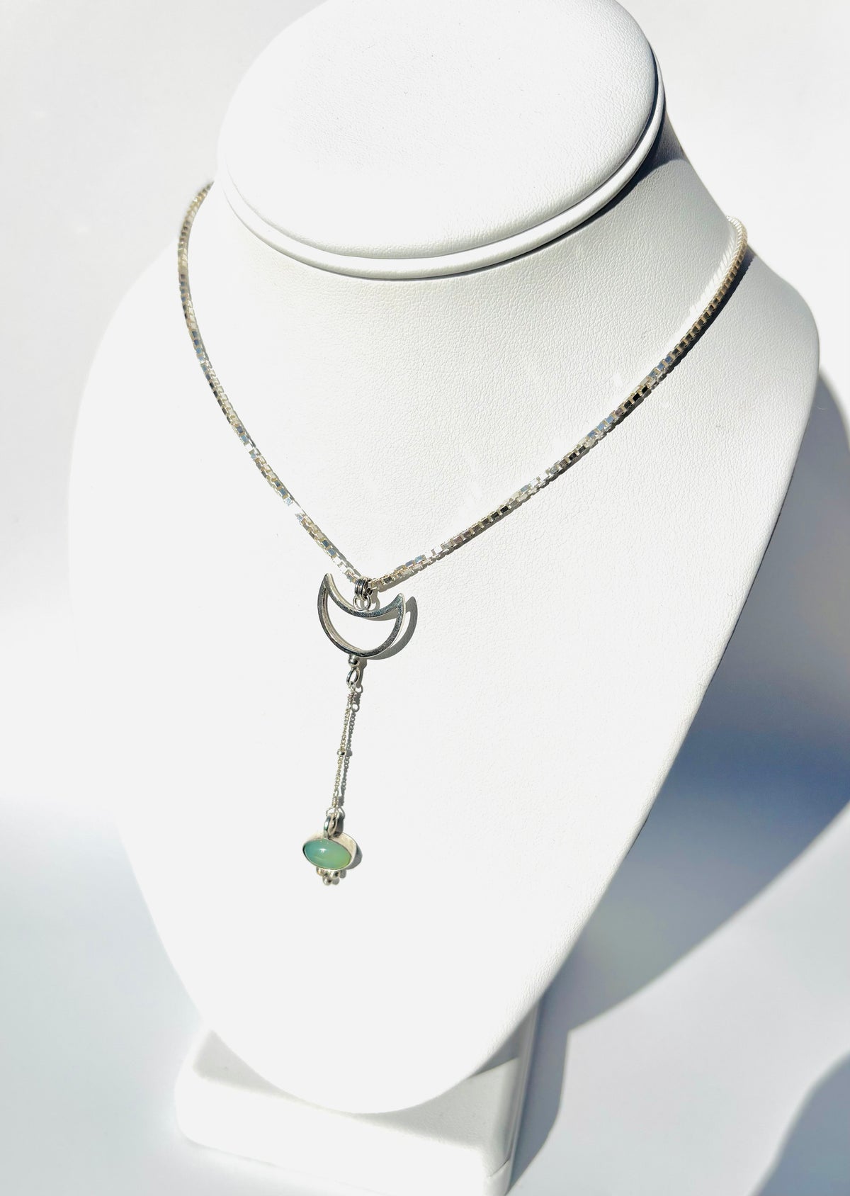 A silver-toned necklace displayed on a white jewelry bust. The necklace features a delicate chain, a crescent moon pendant, and a dangling bar with a small light green heart-shaped stone at the end.