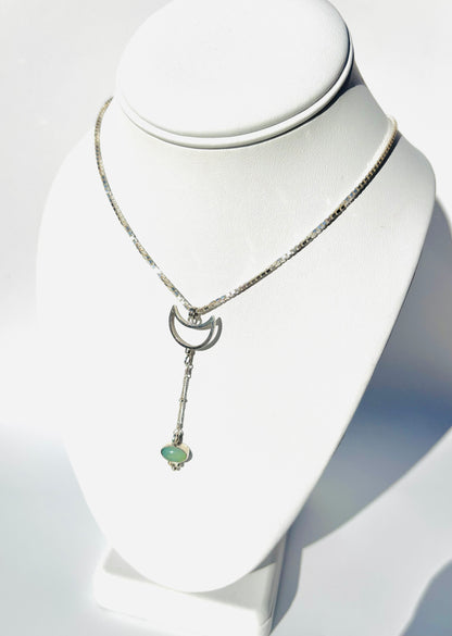 A silver-toned necklace displayed on a white jewelry bust. The necklace features a delicate chain, a crescent moon pendant, and a dangling bar with a small light green heart-shaped stone at the end.